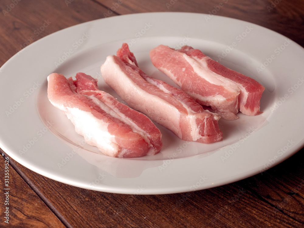 Uncooked raw pork belly slices on a white plate and wooden table. Uncooked raw pork belly slices on a white plate and wooden table.