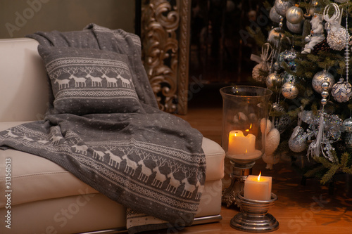Christmas still life with grey nordic knit blanket and cushion with deers pattern on a couch, big candles on a floor and decorated Christmas tree on the back
