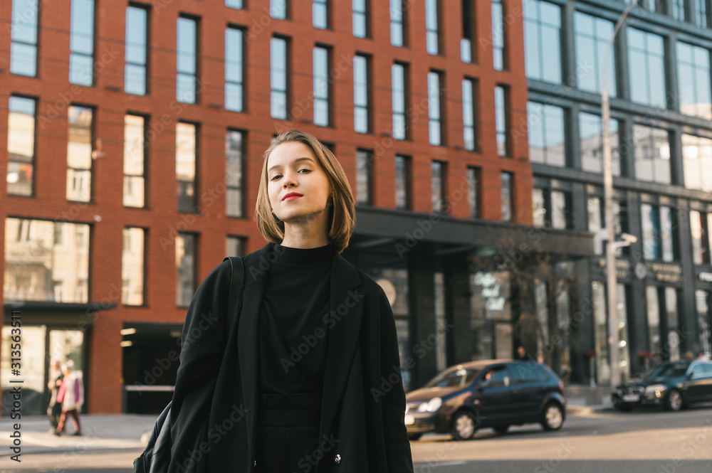 Attractive girl in casual fashion poses against the background of modern architecture and the street, looks into the camera with a smile. Young business lady standing on the street of a megapolis Attractive girl in casual fashion poses against the background of modern architecture and the street, looks into the camera with a smile. Young business lady standing on the street of a megapolis