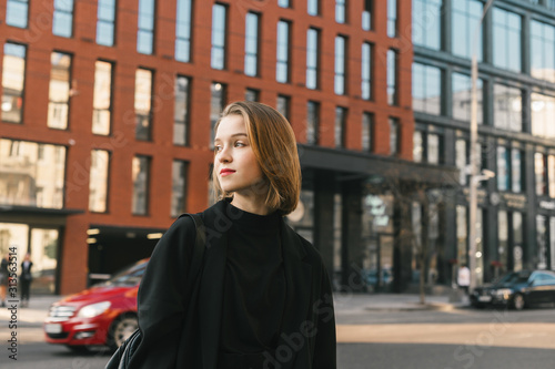 Street portrait of a stylish girl in dark casual clothes stands on the street of a metropolis and a modern building, looking away. Lifestyle photo of a fashionable lady in a jacket and with a backpack