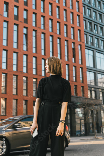 Street portrait of a stylish girl in dark casual clothes stands on the street of a metropolis and a modern building, looking away. Lifestyle photo of a fashionable lady in a jacket and with a backpack