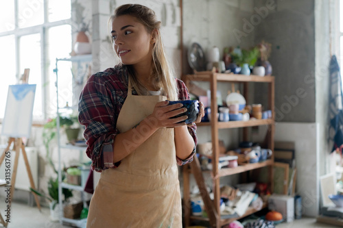 Young lovely smiling girl with blonde hair in apron holding a clay pot in her hands and standing in pottery studio. Pottery workshop. Clay model.