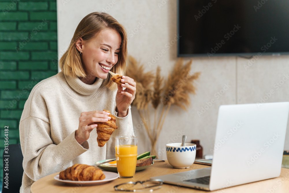 Photo of joyful blonde woman using laptop while having breakfast Photo of joyful blonde woman using laptop while having breakfast