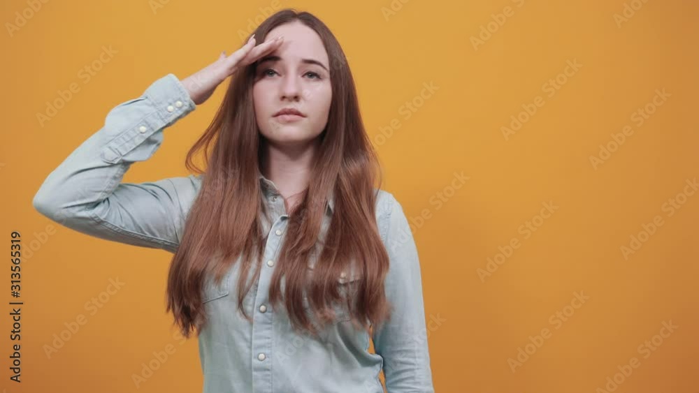 Focused caucasian brunette woman in denim blue shirt keeping palm at forehead, looking into distance isolated on orange background in studio. People emotions, lifestyle concept. Focused caucasian brunette woman in denim blue shirt keeping palm at forehead, looking into distance isolated on orange background in studio. People emotions, lifestyle concept.