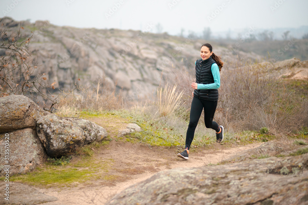 Beautiful Smiling Woman Running on the Mountain Trail at Cold Autumn Evening. Sport and Active Lifestyle. Beautiful Smiling Woman Running on the Mountain Trail at Cold Autumn Evening. Sport and Active Lifestyle.