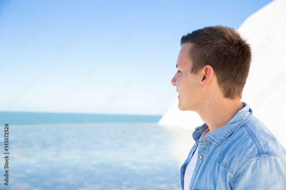 Pensive student guy relaxing at sea, looking at horizon. Handsome young man in casual walking along promenade. Male portrait or advertising concept