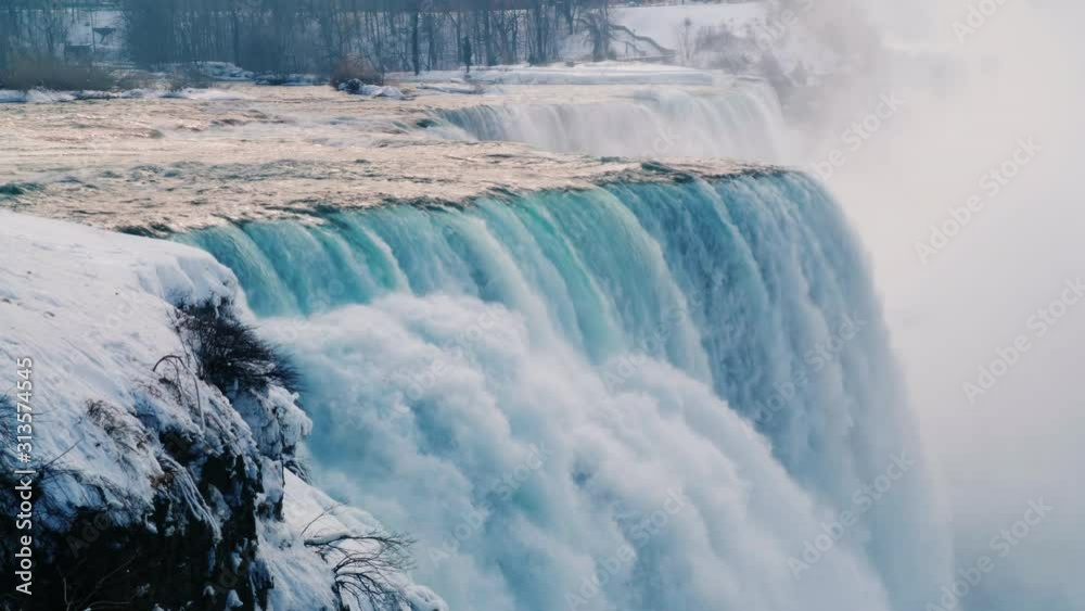 Niagara Falls in winter time. Streams of water flow surrounded by snow-covered shore.