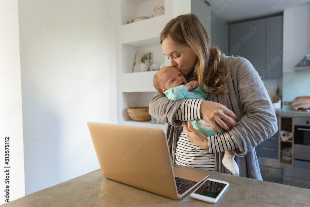 New mother holding and rocking crying baby near laptop. Portrait of young woman and cute little child in home interior. Communication concept New mother holding and rocking crying baby near laptop. Portrait of young woman and cute little child in home interior. Communication concept