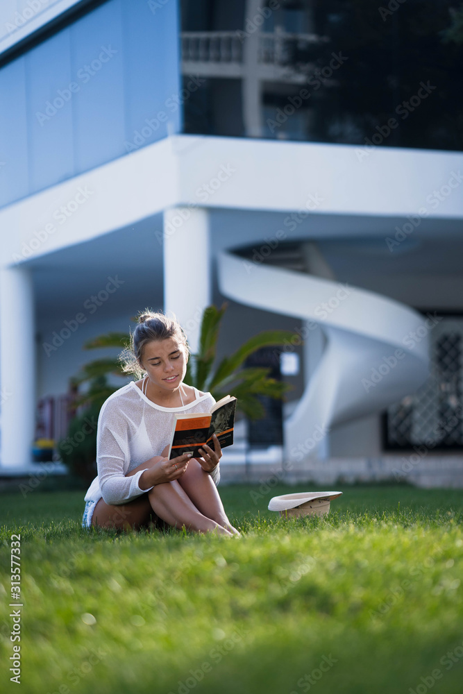 Fototapeta premium Beautiful smart girl with gathered hair sitting on a green lawn and reading book. Girl wearing in bikini and white tunic. The suns rays shining on her.
