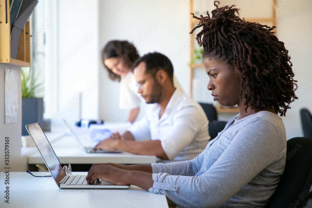 Side view of African American woman typing on laptop. Concentrated colleagues working with modern devices while sitting at table. Technology concept Side view of African American woman typing on laptop. Concentrated colleagues working with modern devices while sitting at table. Technology concept