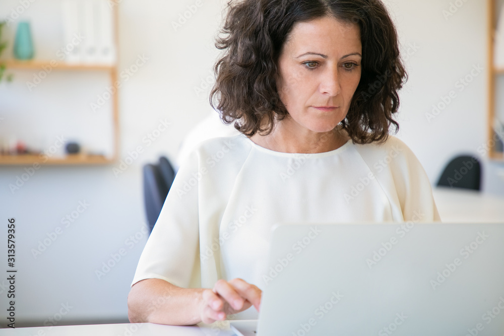 Front view of thoughtful woman using laptop in office. Concentrated mature lady wearing white blouse working with laptop. Technology concept Front view of thoughtful woman using laptop in office. Concentrated mature lady wearing white blouse working with laptop. Technology concept