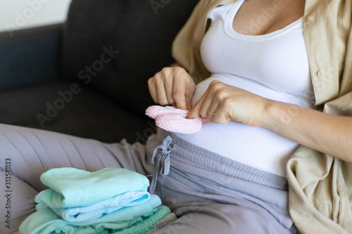 Closeup of expectant mother sitting on couch with garment, applying pink baby shoes to belly. Pregnant young woman spending leisure time at home. Expecting baby girl concept