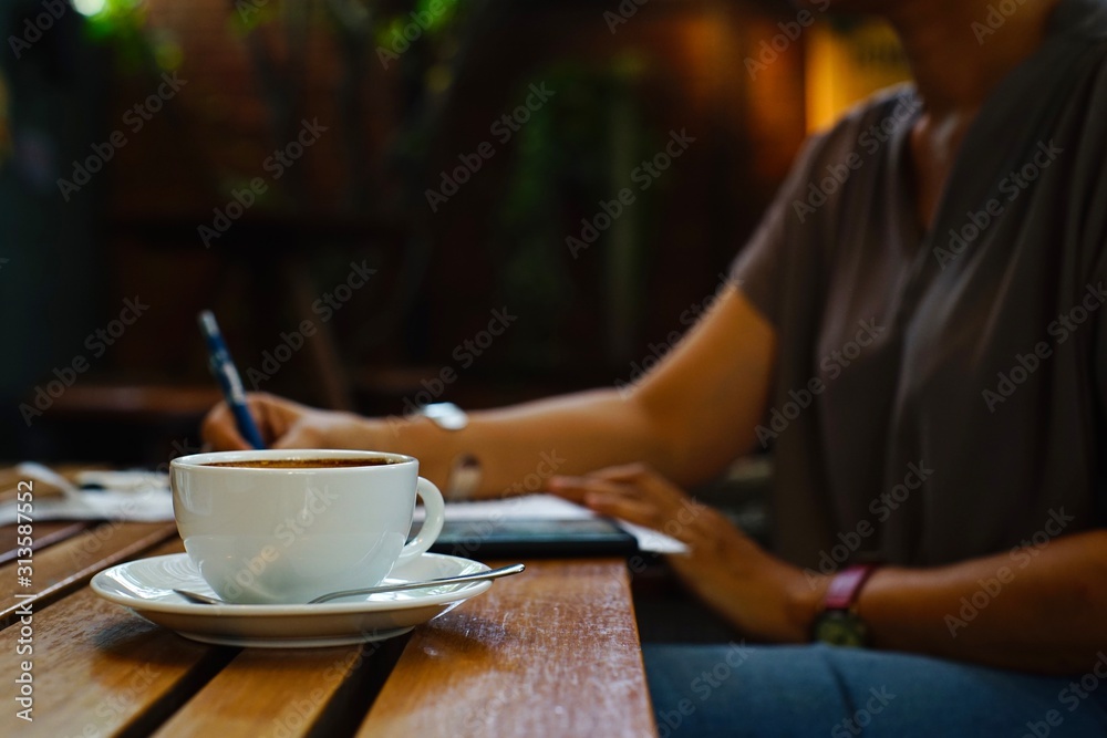Hot coffee in a white mug placed beside the woman who was writing and reviewing documents. Hot coffee in a white mug placed beside the woman who was writing and reviewing documents.