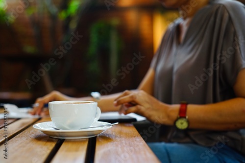 Hot coffee in a white mug is placed on the table in front of a woman's hand typing a message on the mobile phone.