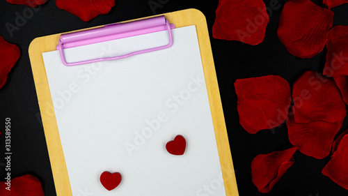 Shot from above on a black background with a clipboard with a white blank sheet on which there are red hearts. Rose petals lay around on a black background. Valentine's Day.
