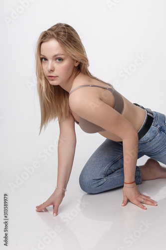 portrait of sexy caucasian woman with long hair posing in beige lingerie and blue jeans on white studio background. model tests of pretty girl in bra. attractive female sitting on floor on her knees