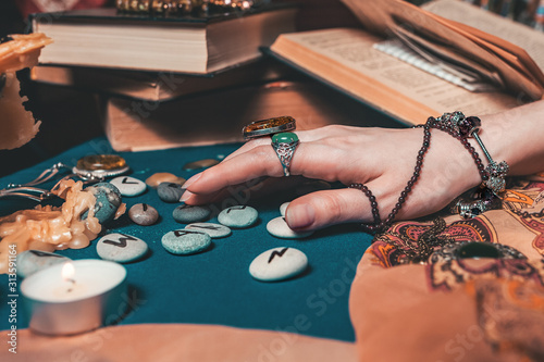 Astrology and esotericism. The female hand of the sorceress reads the runes. In the background, old books, fortune-telling runes, candles, and jewels. Close up