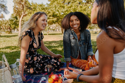 Group of smiling multiracial female best friends sitting together on blanket with fruits enjoying at picnic in the park - group of healthy friends having a picnic