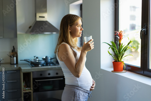 Cuadro en lienzo Side of pensive expectant mother standing in kitchen with glass of water, looking through window