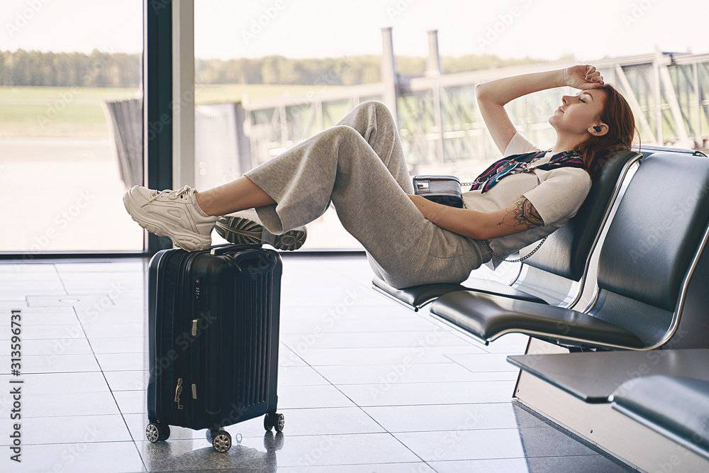 Airport travel lifestyle. Young Gen Z girl traveler sitting in terminal hall with her luggage, trolley bag, passport, smartphone and ticket while waiting for her flight before departure.