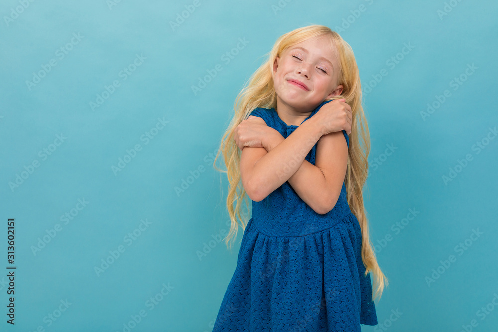 Little girl with blonde hair in blue dress is relaxed and enjoying, portrait isolated on blue background