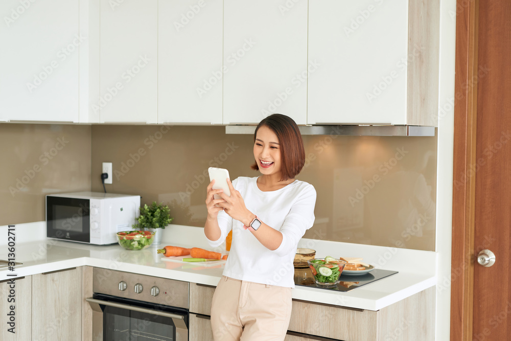 Beautiful young woman standing by kitchen counter reading text message on her cell phone. Beautiful young woman standing by kitchen counter reading text message on her cell phone.