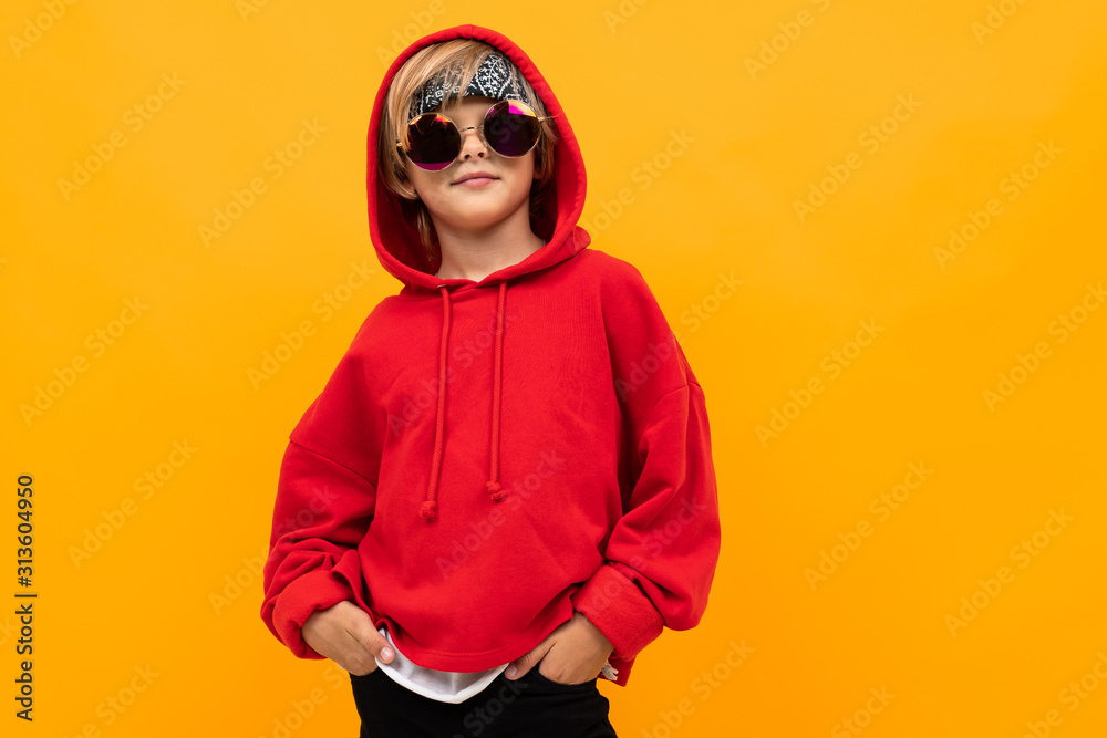 blond boy with a bandana on his head in a red hoodie and glasses posing on an orange background blond boy with a bandana on his head in a red hoodie and glasses posing on an orange background