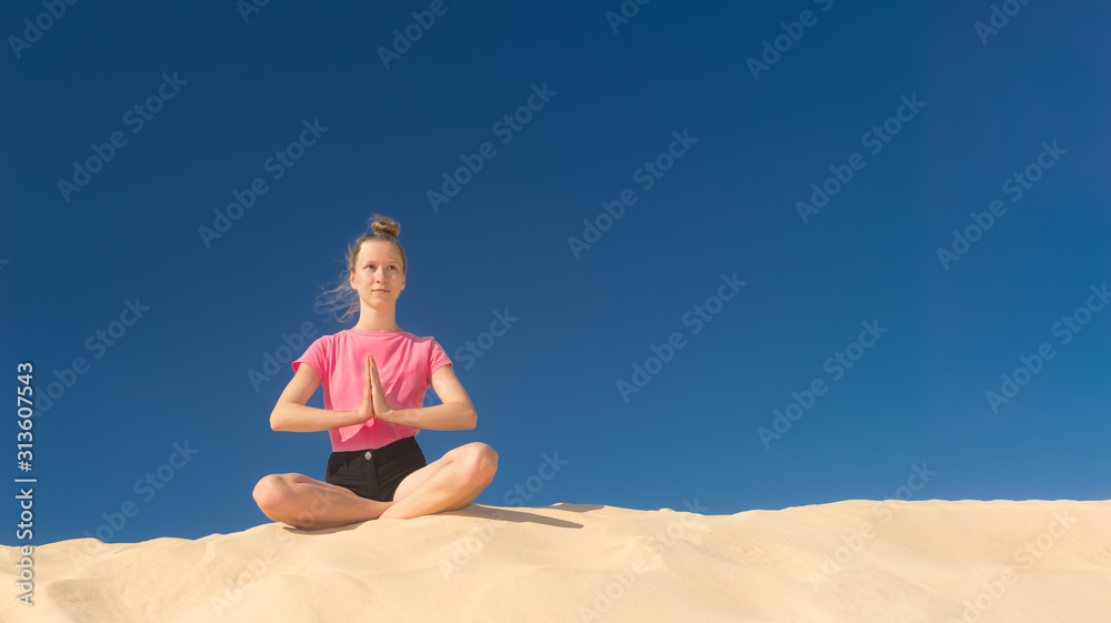 ein junges Mädchen macht im Sommer Yoga auf einer Düne im gelben Sand vor blauem Himmel