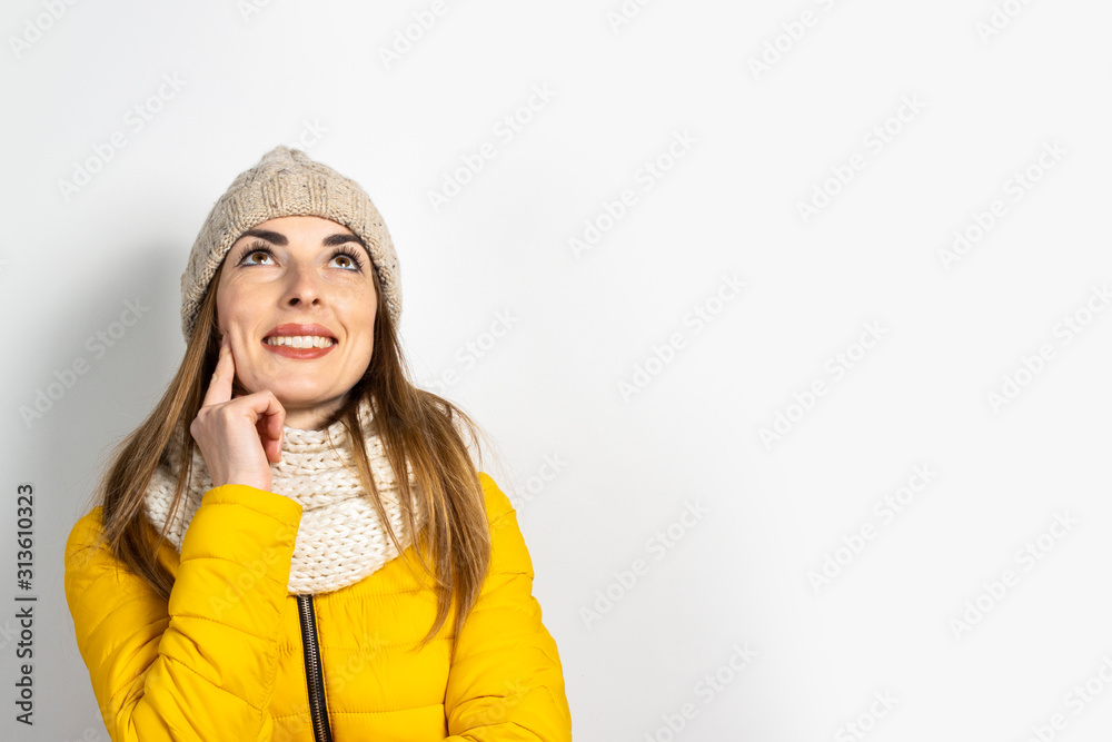 Young woman in a yellow down jacket and hat with a pensive face on a light background. Concept dream, thought, choice, decision, planning. Banner Young woman in a yellow down jacket and hat with a pensive face on a light background. Concept dream, thought, choice, decision, planning. Banner