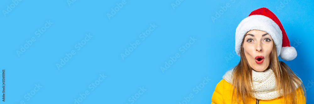 Young woman with a surprised face in a yellow jacket and a hat of Santa Claus on a blue background. Concept of the winter holidays, Christmas, New Year, surprise, shock. Banner