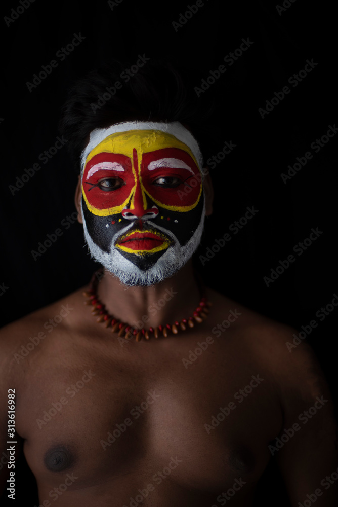 Face of Indian brunette man with his face illuminated and painted by vibrant colors like a tribe standing with candle light in front of a black studio copy space background. Indian hi fashion.