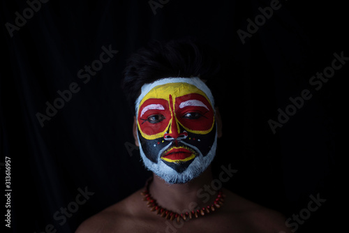 Face of Indian brunette man with his face illuminated and painted by vibrant colors like a tribe standing with candle light in front of a black studio copy space background. Indian hi fashion.