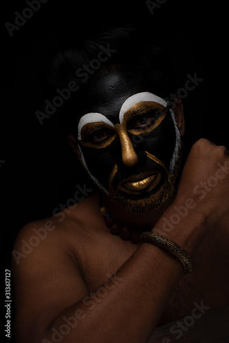 Close up of Indian brunette man with his face illuminated and painted by black and golden like a tribe standing with candle light in front of a black studio copy space background. Indian hi fashion.