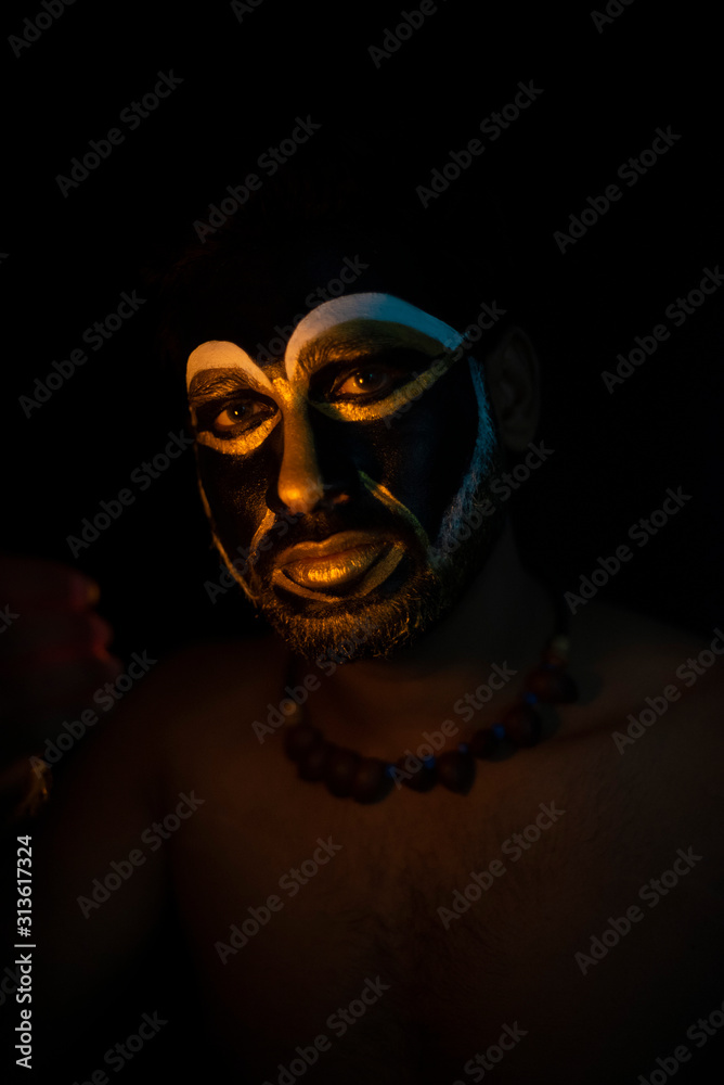 Close up of Indian brunette man with his face illuminated and painted by black and golden like a tribe standing with candle light in front of a black studio copy space background. Indian hi fashion. Close up of Indian brunette man with his face illuminated and painted by black and golden like a tribe standing with candle light in front of a black studio copy space background. Indian hi fashion.