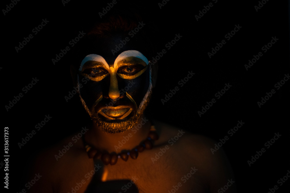 Close up of Indian brunette man with his face illuminated and painted by black and golden like a tribe standing with candle light in front of a black studio copy space background. Indian hi fashion. Close up of Indian brunette man with his face illuminated and painted by black and golden like a tribe standing with candle light in front of a black studio copy space background. Indian hi fashion.
