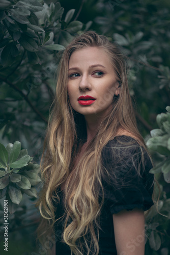 Blond girl wearing red lipstick, has long hair and  looking back. Forest. Green. 