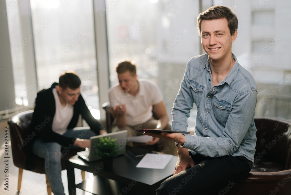 Focused young man uses a tablet and looking at the camera in sunlit office. Two young startup businessman work together behind a laptop in the background. Focused young man uses a tablet and looking at the camera in sunlit office. Two young startup businessman work together behind a laptop in the background.