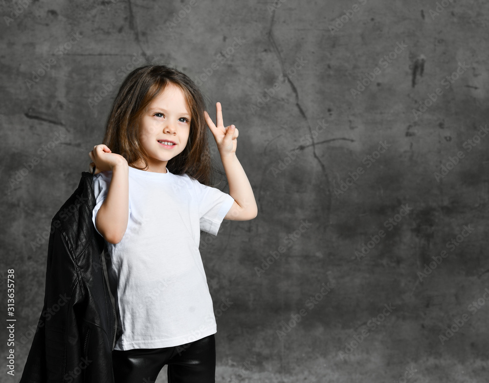 Small smiling cute girl in black and white rock style clothing standing and showing peace and victory sign with fingers Small smiling cute girl in black and white rock style clothing standing and showing peace and victory sign with fingers