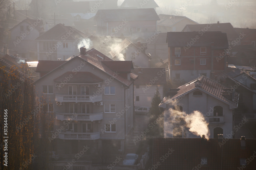 Naklejka Dymiące kominy na dachach domów wydzielają dym, smog o wschodzie słońca, zanieczyszczenia przedostają się do atmosfery. Katastrofa ekologiczna. Szkodliwe emisje i spaliny do powietrza. Mgła, zimowy dzień, sezon grzewczy.