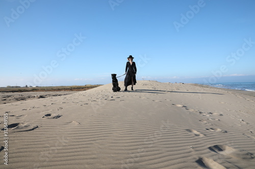 A woman standing on top of a sandy beach with dog. Outdoor.