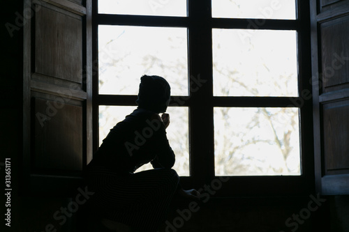Silueta de mujer pensativa delante de un ventanal con gorro, confinación.