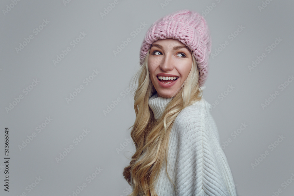 Happy young woman with long blond hair in pink knitted winter hat looking back over her shoulder with a beaming smile against a grey background Happy young woman with long blond hair in pink knitted winter hat looking back over her shoulder with a beaming smile against a grey background