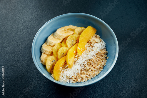 Close up view on Healthy breakfast: oatmeal with coconut, mango, bananas and yellow kiwi in a blue bowl on a black background.