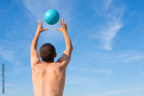 A man with a ball against a blue sky plays beach volleyball. The view from the back. Life style.