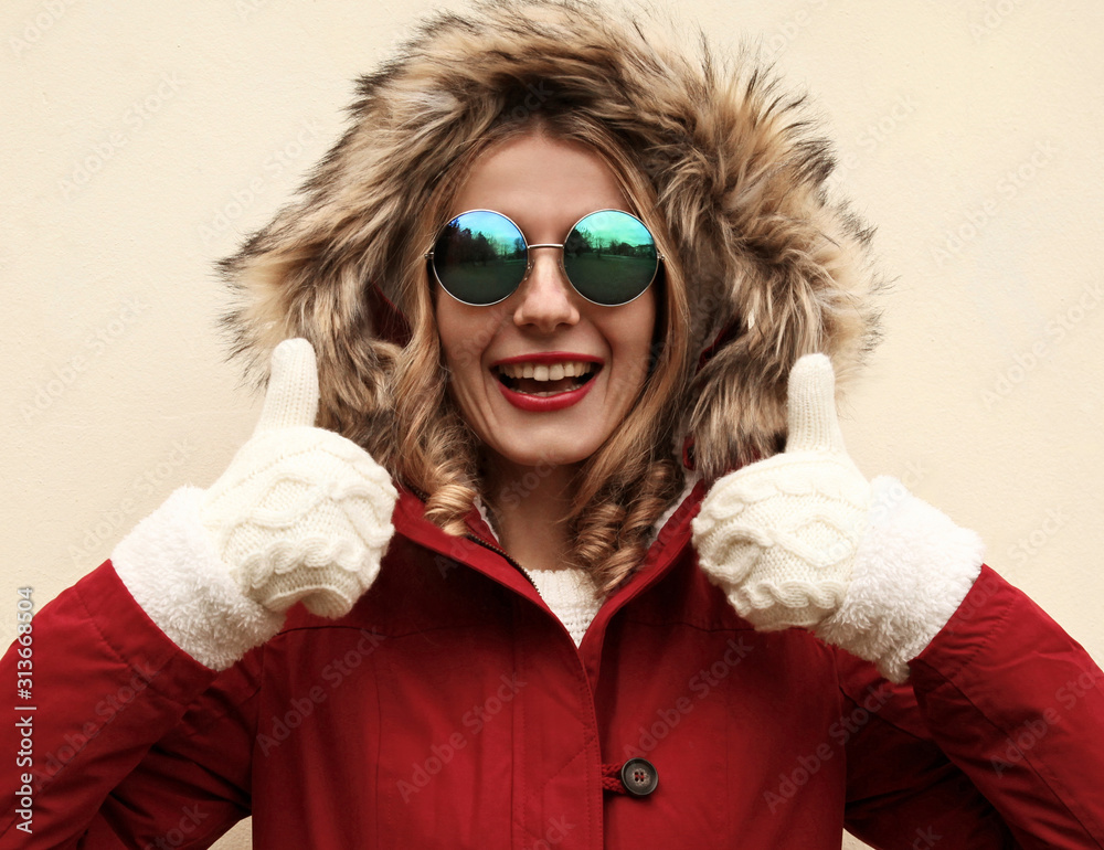 Portrait close up happy smiling woman showing thumbs up as like sign wearing red jacket with fur hood and white mittens over background Portrait close up happy smiling woman showing thumbs up as like sign wearing red jacket with fur hood and white mittens over background