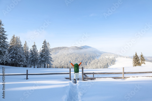 Winter landscape. On the path near the wooden fence stands a girl with a tourist backpack. The meadow overlooks the mountains, the forest with trees covered with snow and the blue sky.