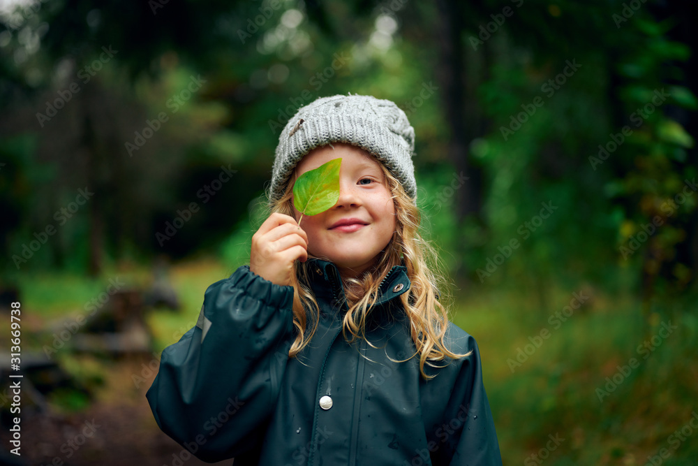 Funny girl in warm hat closing eyes with green leaf in forest Funny girl in warm hat closing eyes with green leaf in forest