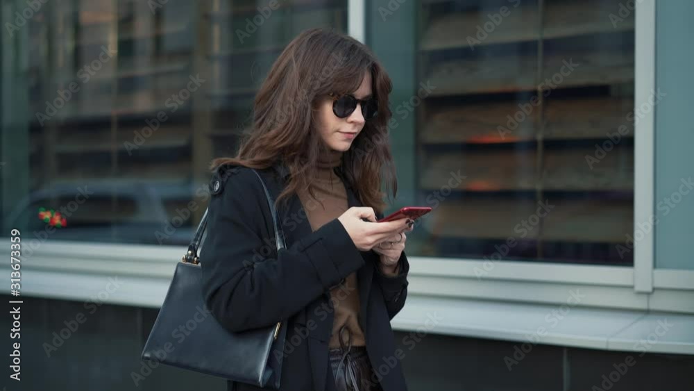 Slow motion of woman in sunglasses with a red phone and a bag on background of shop windows in a big city. Gimbal shot of woman in black coat typing in the phone looking to the road Slow motion of woman in sunglasses with a red phone and a bag on background of shop windows in a big city. Gimbal shot of woman in black coat typing in the phone looking to the road