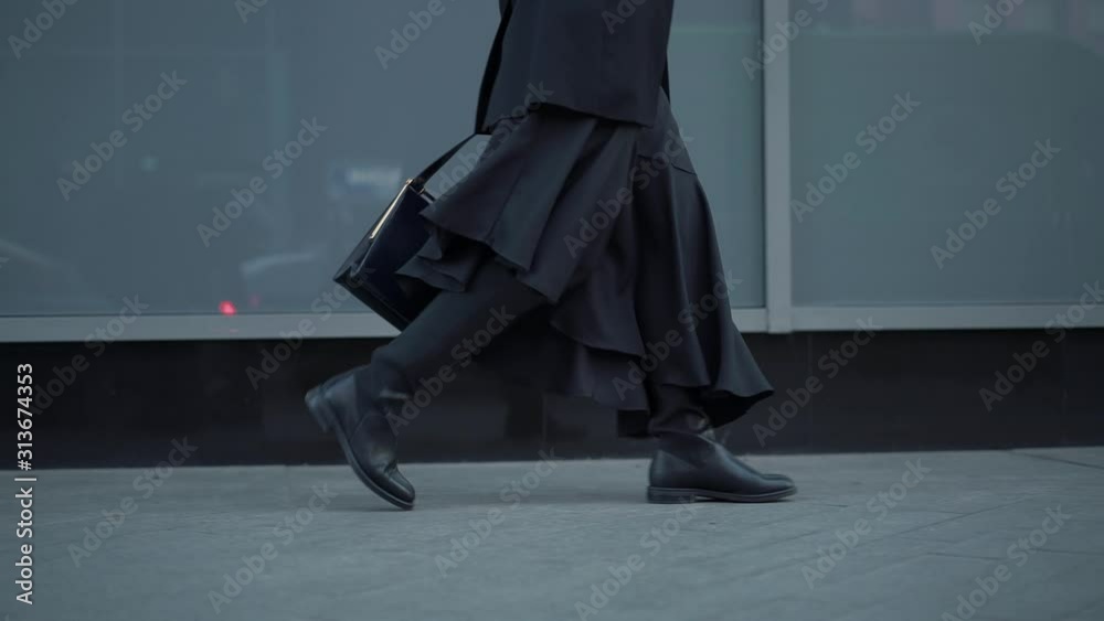 Slow motion gimbal shot of woman dressed in black walking from right to left in black boots on the city pavement on the background of grey shop window glasses
