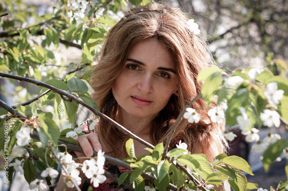 soft focus of beautiful woman with long hair standing between blooming branches with white flowers and green leaves and looking at camera on backyard soft focus of beautiful woman with long hair standing between blooming branches with white flowers and green leaves and looking at camera on backyard